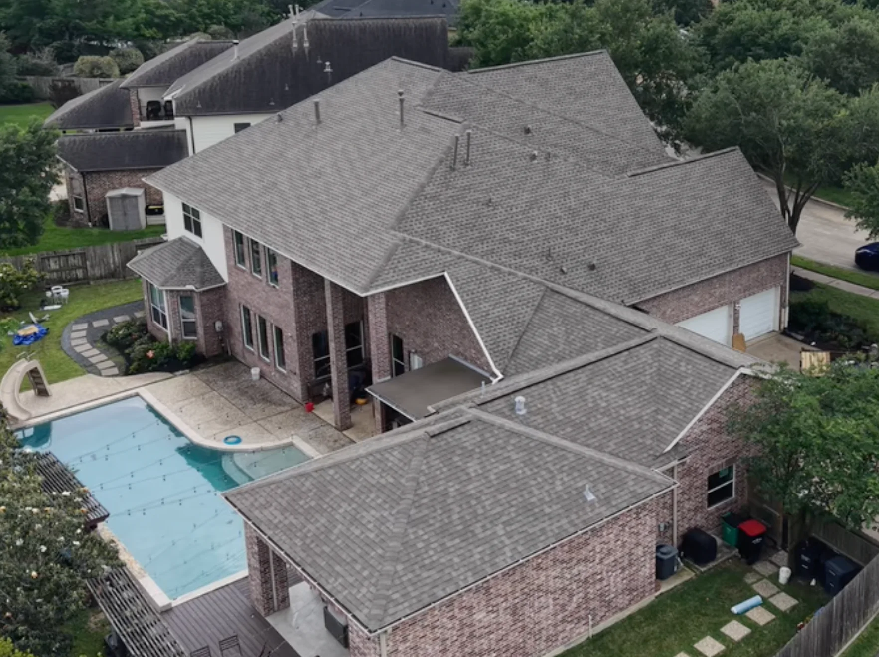 Aerial view of a dark shingle roof on a large brick home with a pool.