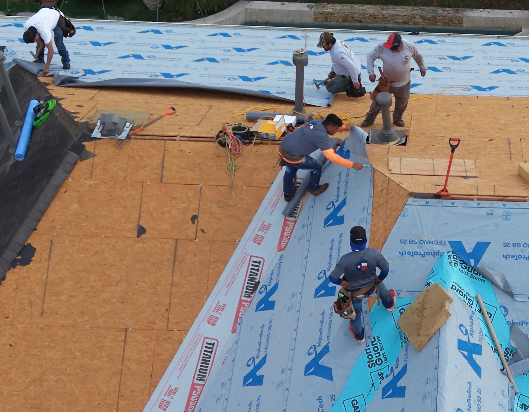Closer aerial view of a roofing crew working across exposed decking and blue underlayment.