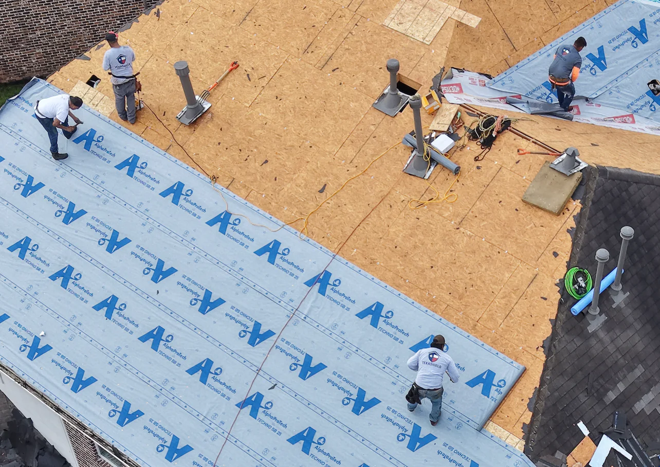 Wide aerial view of a roofing crew inspecting decking and placing blue underlayment.